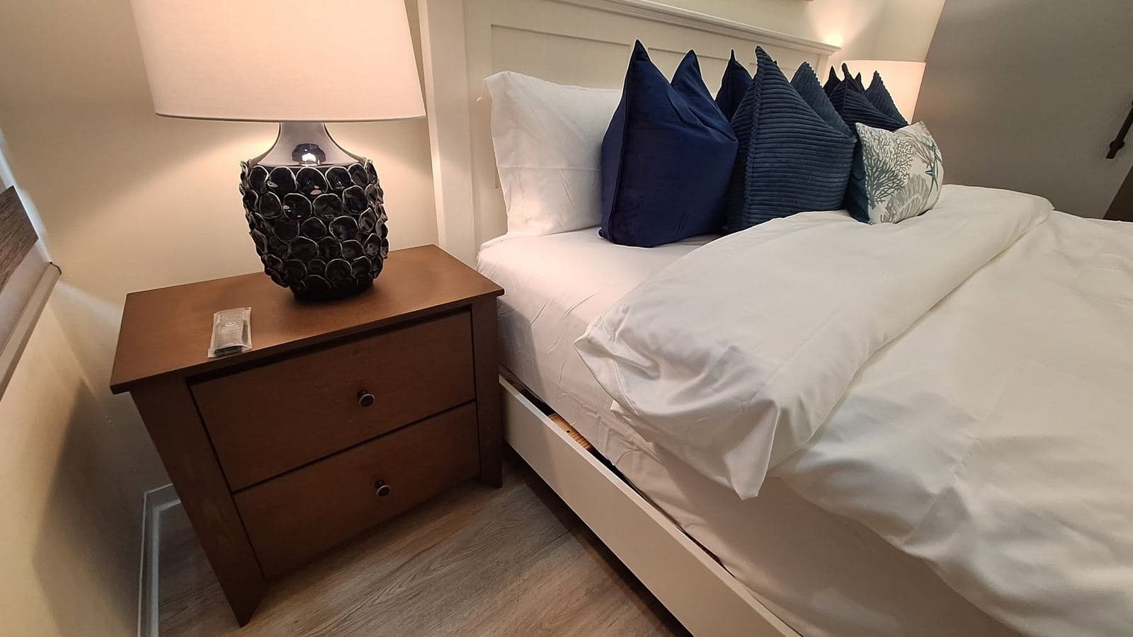 Walnut two-drawer nightstand beside a white panel bed, lit by a warm table lamp